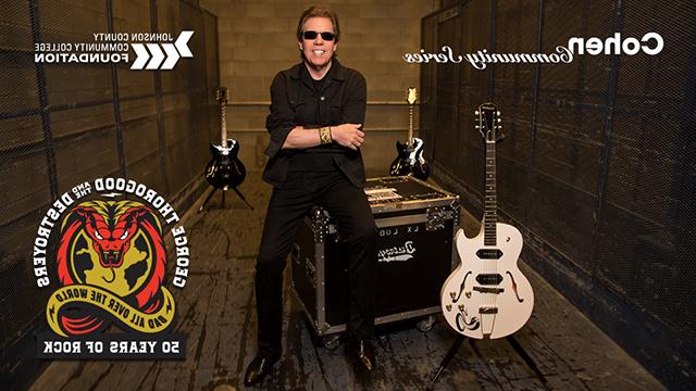 George Thorogood sits next to his guitar on an amp. He has his arms crossed, is smiling at the camera and is wearing sunglasses.