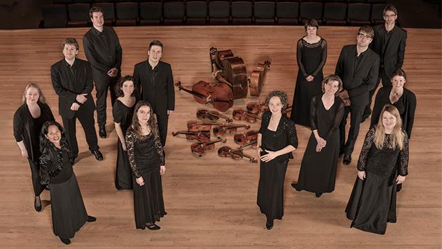 The 14 members of Les Violons pose for a picture. The picture shows them from over head standing in two groups on stage with their violins arranged on the floor in the middle of the stage.
