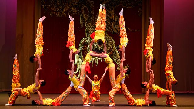 a group of 16 acrobats perform a stunt on stage where seven of them are doing hand-stands with the other acrobats as bases